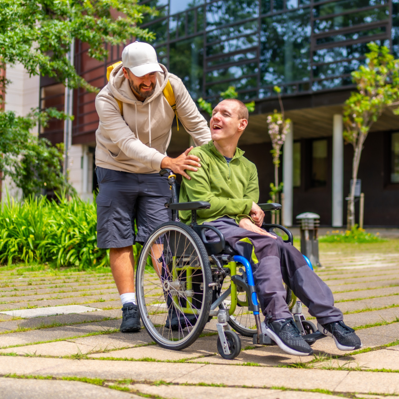 Helfer schiebt Mann im Rollstuhl über gepflasterten Weg
