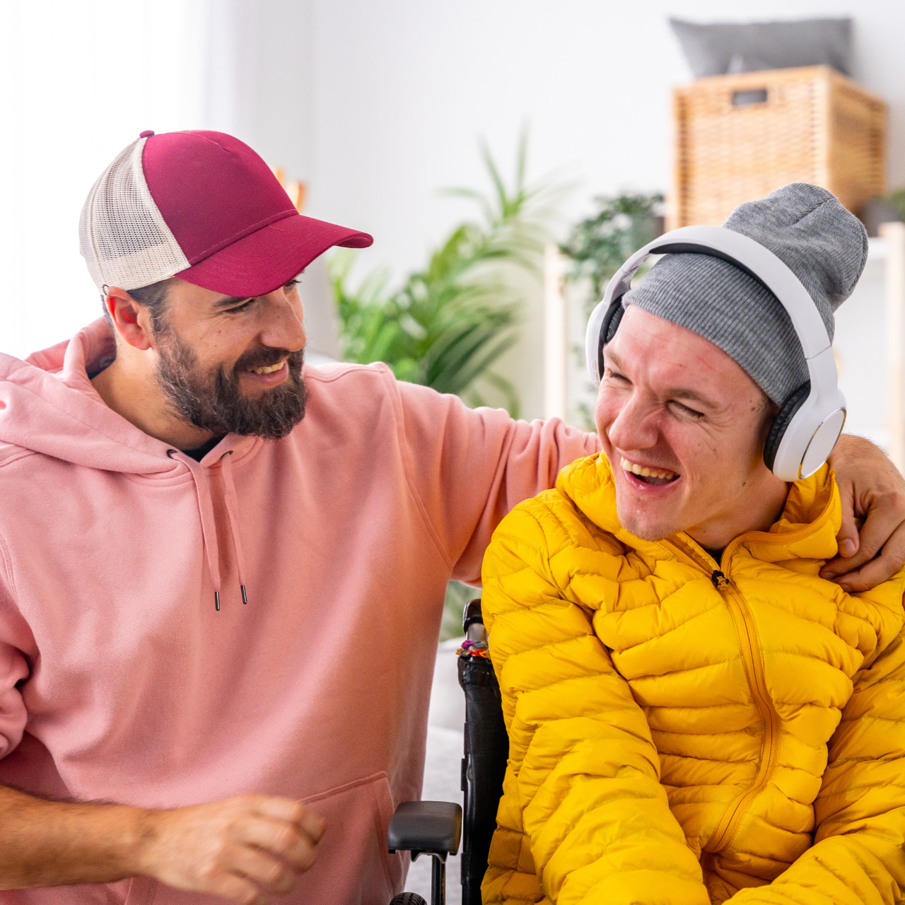Zwei Personen lachen sich an. Eine davon sitzt im Rollstuhl und trägt Kopfhörer