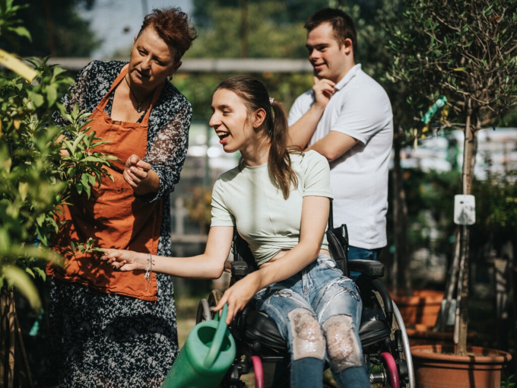 Drei Personen schauen auf einen Baum. Frau im Rollstuhl hat Gießkanne in der Hand