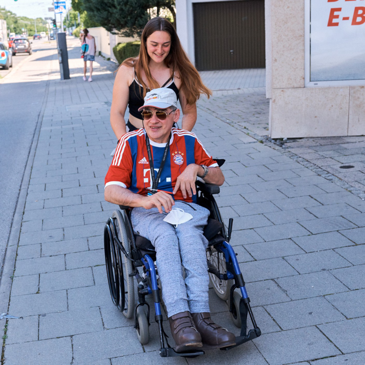 Helferin schiebt Mann im Rollstuhl mit Bayerntrikot auf Gehweg
